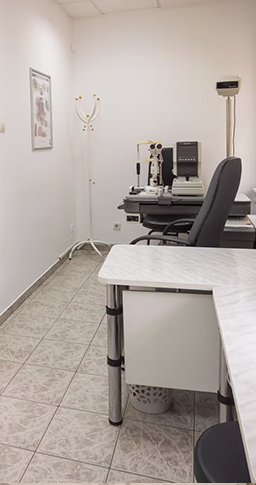 На фотографии изображён кабинет офтальмолога. В конце кабинета стоит аппарат для проверки зрения. Рядом с ним находиться стол офтальмолога