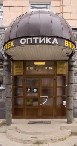 На фотографии изображен вход в магазин Brotex. Фасад входа слегка закруглен. Между дверью стоят две каменные колонны. Над входом изображен логотип Brotex бело-золотого цвета